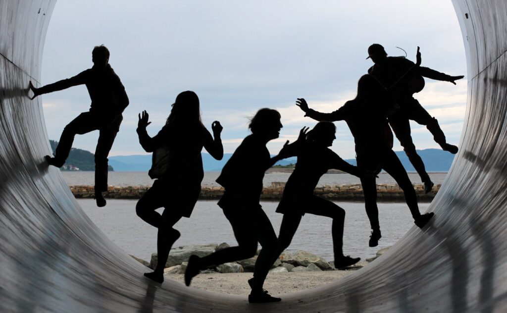 Six people in silhouette climb and jump in a giant silver tube with a beachfront view behind them