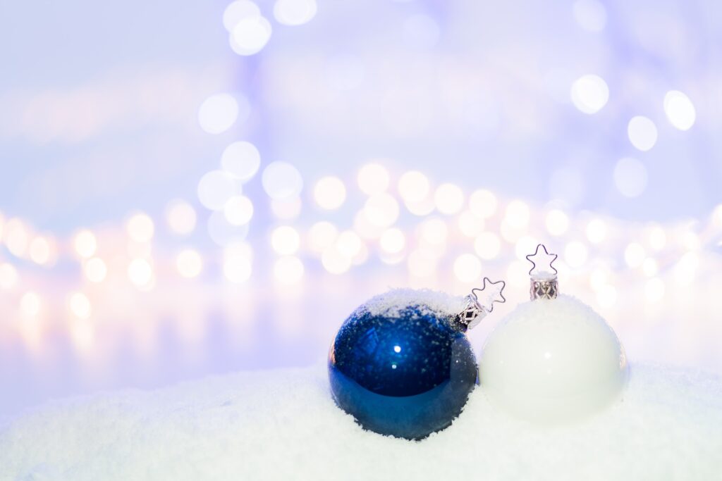 two baubles—one blue, one white—sit in the snow against a light blue background with blurry white lights